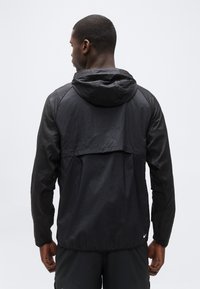 Άνδρας που στέκεται με την πλάτη, φορώντας μια μαύρη ελαφριά ζακέτα με κουκούλα και μαύρο σορτς, σε έναν απλό φόντο.