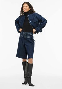 Denimjakk lühendatud lõikega, kantud üle musta kaelusega sviitri. Sobitatud tumedate denimlühikeste pükstega, millel on vöödiga talje ja mustade põlvekõrguste saapadega.