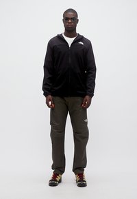 Άνδρας που φοράει μαύρο μπουφάν με φερμουάρ, ελιά παντελόνι, παπούτσια πεζοπορίας και γυαλιά ηλίου, στέκεται μπροστά σε απλό λευκό φόντο.