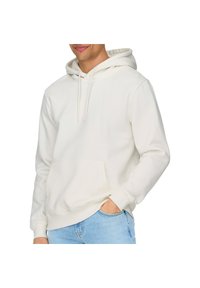 Haut à capuche blanc en matériau doux, avec une poche avant, des cordons de serrage et des poignets côtelés. Associé à un jean bleu clair.
