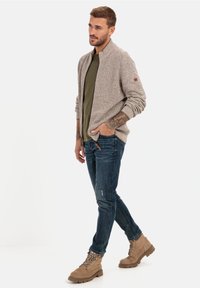 Beige Strickjacke mit hohem Kragen, getragen über einem grünen T-Shirt, kombiniert mit blauen Jeans und tanfarbenen Wanderschuhen mit schwarzen Schnürsenkeln.