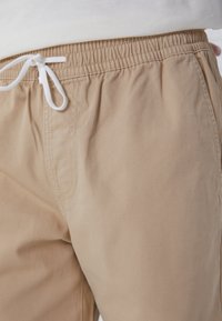 Pantaloni di cotone beige con una vita elastica e un laccio bianco, tasca frontale e una texture liscia, con dettagli di cucitura sottili.