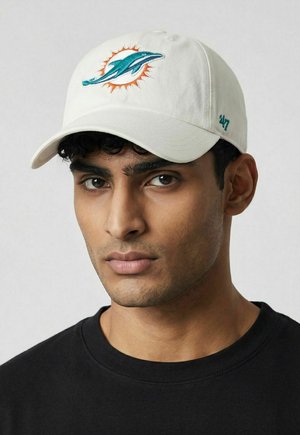 Junger Mann, der eine weiße Kappe mit einem türkisfarbenen Delfin- und orangen Sonnenlogo sowie ein schwarzes Shirt trägt und leicht zur Seite schaut.