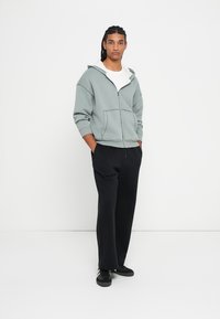 Light gray zip-up hoodie with a kangaroo pocket, paired with black trousers and black sneakers, featuring a relaxed fit and cotton texture.