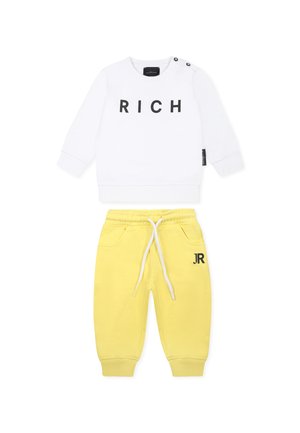 Biała bluza z czarnym napisem "RICH" i szczegółami w postaci guzików po bokach, połączona z żółtymi spodniami dresowymi z elastycznym pasem i logo "JR".