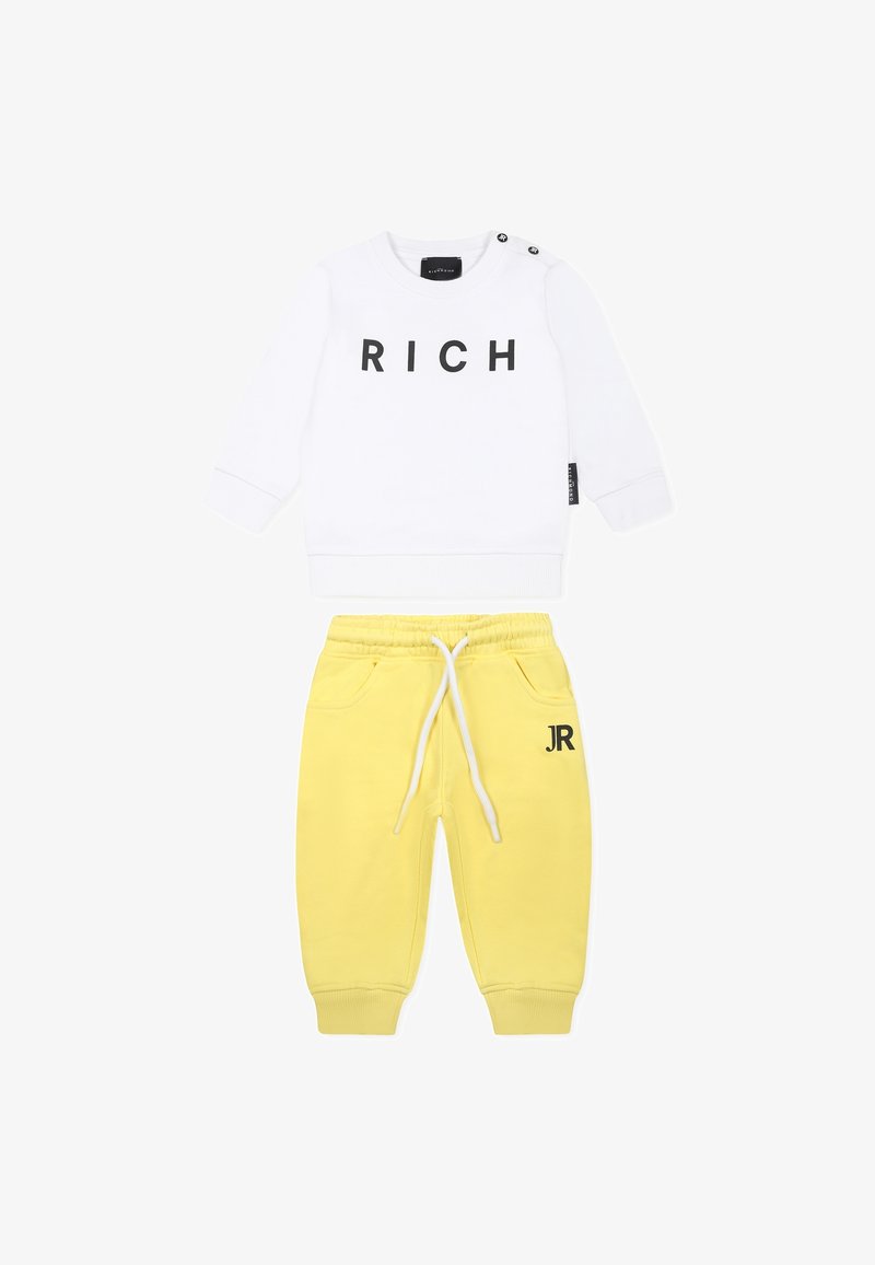 Biała bluza z czarnym napisem "RICH" i szczegółami w postaci guzików po bokach, połączona z żółtymi spodniami dresowymi z elastycznym pasem i logo "JR".