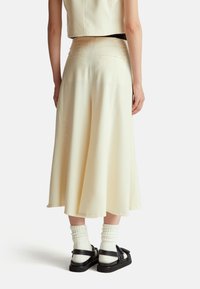 Světle žlutá midi sukně vyrobená z hladkého materiálu s rozšířeným střihem a bočními kapsami. Nosená s černými sandály a bílými žebrovanými ponožkami.