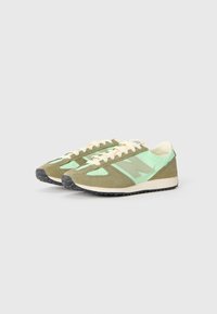 Une paire de baskets New Balance en suède vert clair et olive avec des lacets blancs et des semelles en caoutchouc noir sur un fond uni.
