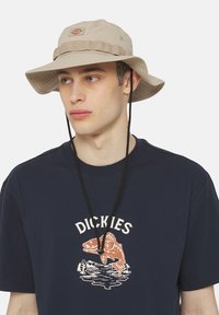 Beige bøttehatt med en teksturert band, justerbar hake-rem og logo. Brukt med en marineblå sweatshirt med et fiske-graphic.