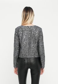 Geknipte grijze paillettenjasje met lange mouwen, met een algeheel patroon van pailletten in verschillende maten. Gegd gedragen met strakke zwarte broek.