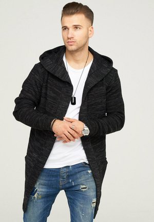 Black knitted long cardigan with a hood, open front, and two pockets, worn over a white t-shirt and distressed blue jeans.