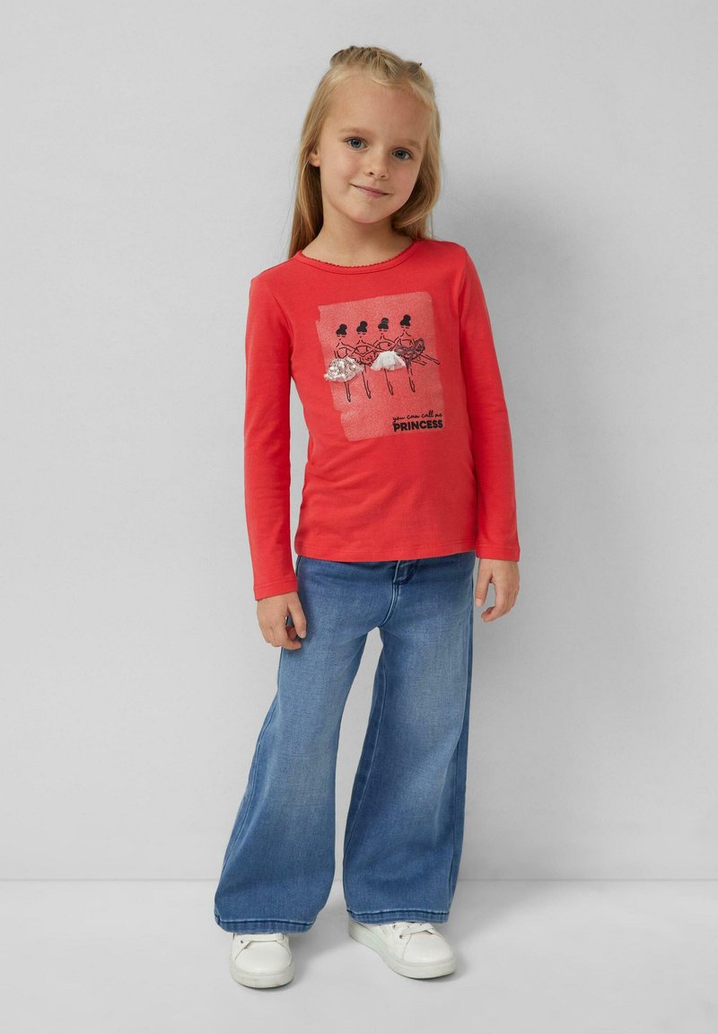 Rotes Langarmshirt mit einem Druckdesign von Tänzern und der Aufschrift "Du kannst mich Prinzessin nennen." Kombiniert mit hellblauen Schlagjeans und weißen Schuhen.