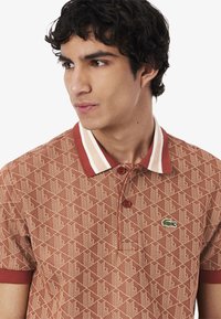 Kortärmad polo i rostfärg med geometriskt mönster, kontrasterande krage med krämfärgade och burgundy ränder samt en liten grön logga.