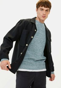 Black button-up jacket with two front pockets, worn over a textured gray sweater. Model has short hair and blue eyes.