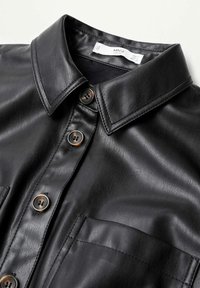 Chemise noire en cuir synthétique avec col pointu, quatre boutons et une poche poitrine, posée à plat sur une surface blanche.