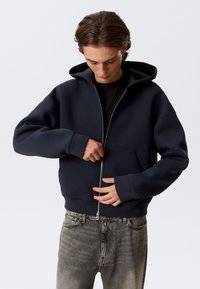 Sweat à capuche zippé bleu marine en matière douce avec une grande capuche, une poche avant et des poignets côtelés, assorti à un jean gris clair.