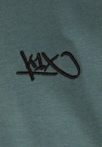 Bavlněné tričko v tlumené modrozelné barvě s černým vyšívaným logem, které obsahuje stylizovaný text a linie. Hladká textura, minimalistický design.