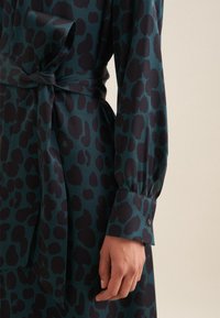 Leopardenmuster-Kleid aus dunkelgrünem und schwarzem Stoff, mit gebundenem Taillenbund, langen puffärmeln und knopfepi. 