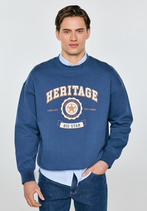 Młody mężczyzna w niebieskiej bluzie z napisem "HERITAGE BIG STAR", jasnoniebieskiej koszuli w paski i niebieskich dżinsach, stojący na jednolitym tle.