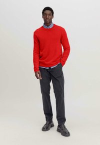 Roter gestrickter Pullover mit Rundhalsausschnitt, über einem hellblauen Hemd mit Kragen getragen. Kombiniert mit dunkelgrauen Hosen und schwarzen Slippern.