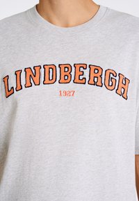 Grå kortärmad t-shirt gjord av mjukt tyg. Har svart och orange broderad text "LINDBERGH" och "1927" nedanför. Enkel design.