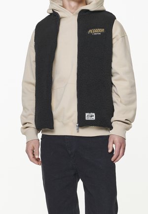 Mand iført beige hættetrøje og sort fleecevest med lynlås foran med "Pegador London" logo, kombineret med sorte bukser, stående mod en hvid baggrund.