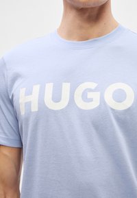 Ανοιχτό μπλε βαμβακερό T-shirt με μεγάλη, έντονη λευκή γραμματοσειρά που γράφει "HUGO." Πρότυπος στρογγυλός λαιμός και κοντά μανίκια. Λείος υφή.