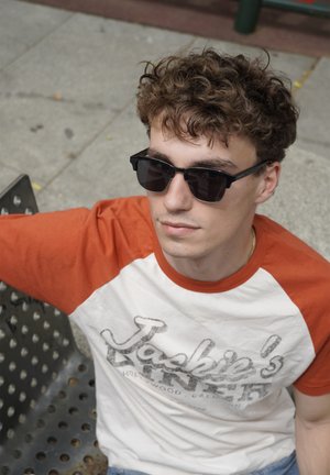 Black sunglasses with square frames, paired with a red and white retro T-shirt. Sitting on a textured gray surface.