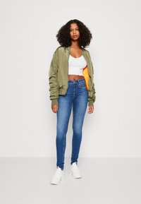 Grønn bomberjakke med oransje fôr, hvit cropped topp, blå skinny jeans og hvite sneakers, med glatte teksturer og en tettsittende design.