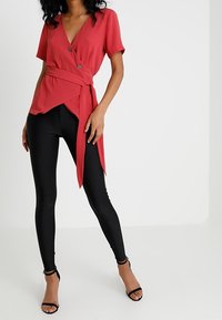 Haut cache-cœur rouge à manches courtes avec un détail noué ; associé à un legging noir ; comporte deux boutons à l'avant et un décolleté en V.