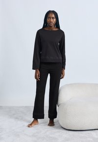 Črna majica z dolgimi rokavi in ujemajoče se hlače s subtilnimi detajli na robu. Mehka, gladka tekstura, sproščen kroj. Model stoji boso.