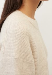 Suéter de lana beige con una superficie suave y texturizada. Presenta un cuello redondo y detalles acanalados a lo largo de los hombros y las costuras laterales.
