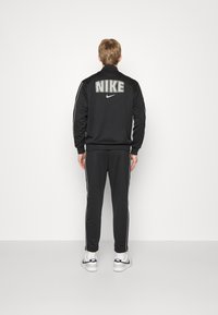 Osoba v černém trenýrkovém kompletu Nike s bílými detaily, stojící čelem k čisté bílé zdi, s velkým logem Nike na zadní části bundy.