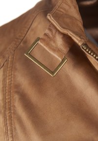 Detail einer braunen Lederjacke mit einer quadratischen Messingschnalle und einer strukturierten Oberfläche mit Nähakzenten. Nahaufnahme betont die Handwerkskunst.