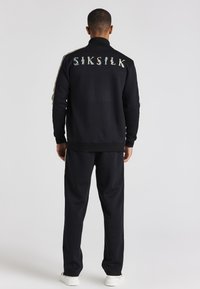 Conjunto de chándal negro que incluye una chaqueta con cremallera y el logotipo bordo "SIKSILK" en la parte posterior, pantalones ajustados y zapatillas blancas. Rayas doradas en los laterales.