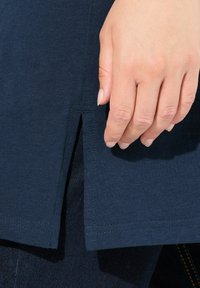 Hemd aus dunkelblauem Baumwollstoff mit seitlichem Schlitz. Nahaufnahme einer Hand, die auf dem Stoff ruht und die glatte Textur hervorhebt.