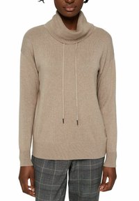 Pull beige à col bénitier avec cordons, poignets et ourlet côtelés. Texture douce, coupe décontractée, associé à un pantalon gris à motifs.
