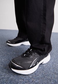 Baskets de sport noires avec tige en maille, semelle blanche et accents noirs. Lacets et surface texturée visibles. Portées avec un pantalon noir sur un tapis.