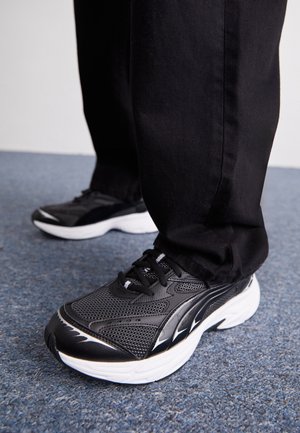 Baskets de sport noires avec tige en maille, semelle blanche et accents noirs. Lacets et surface texturée visibles. Portées avec un pantalon noir sur un tapis.