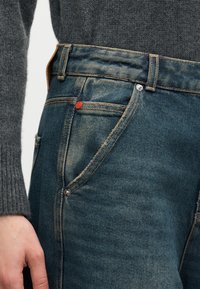 Κοντινό πλάνο ενός ατόμου που φοράει ξεβαμμένα μπλε τζιν και ένα σκούρο γκρι πλεκτό πουλόβερ, δείχνοντας τη μέση και το χέρι που ακουμπά δίπλα στον γοφό.