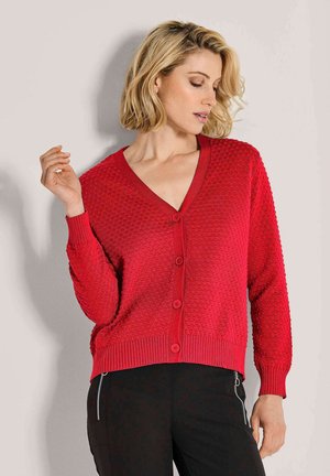 Vrouw met kort blond haar, gekleed in een gestructureerde rode knooptrui en zwarte broek met ritsdetails, poseert tegen een effen lichte muur.