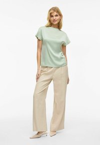 Top in raso verde chiaro con maniche corte, abbinato a pantaloni beige a gamba larga e scarpe con punta in tonalità chiara. Texture liscia.