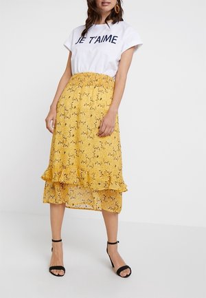 Jupe midi florale jaune avec ourlet en volants, en tissu léger et avec une taille élastique. Associée à un t-shirt graphique blanc et des sandales noires.