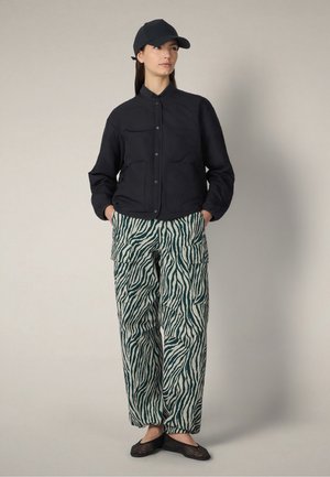 Donna con cappellino nero, giacca nera con bottoni, pantaloni cargo a righe zebrate e scarpe slip-on nere intrecciate, in piedi con le mani nelle tasche.