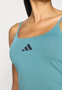 Een turquoise sporttop van gladde stof, met een zwart drie-strepen logo aan de voorkant en dunne schouderbandjes.