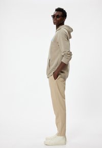 Beige hættetrøje med frontlomme, turkise branding, beige bukser og hvide højtop sneakers. Model iført solbriller, stående sidelæns.