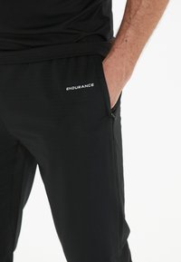 Sorte atletiske bukser lavet af letvægtsmateriale, med en elastisk talje, lynlåslommer og en hvid "ENDURANCE" logo.