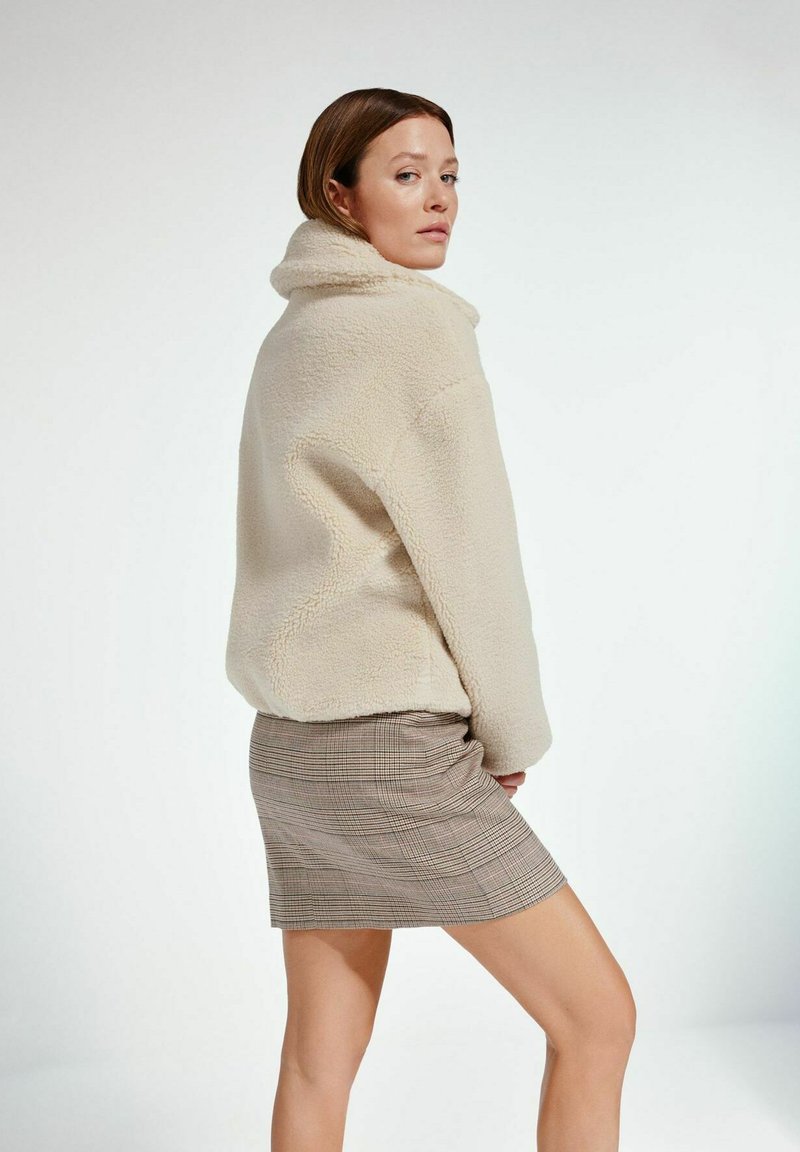 Kremowa polarowa kurtka z wysokim kołnierzem, połączona z spódnicą w kratę. Kurtka ma teksturowany, miękki materiał.