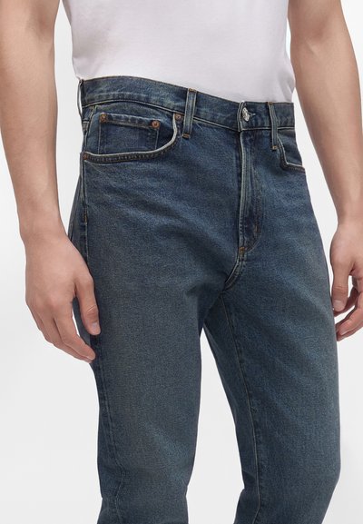 Homme portant un jean en denim bleu foncé et un t-shirt blanc, mains détendues sur les côtés, debout devant un fond uni.