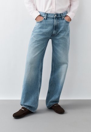 Personne portant un jean large bleu clair, une chemise blanche boutonnée et des mocassins marron foncé, debout les mains dans les poches.